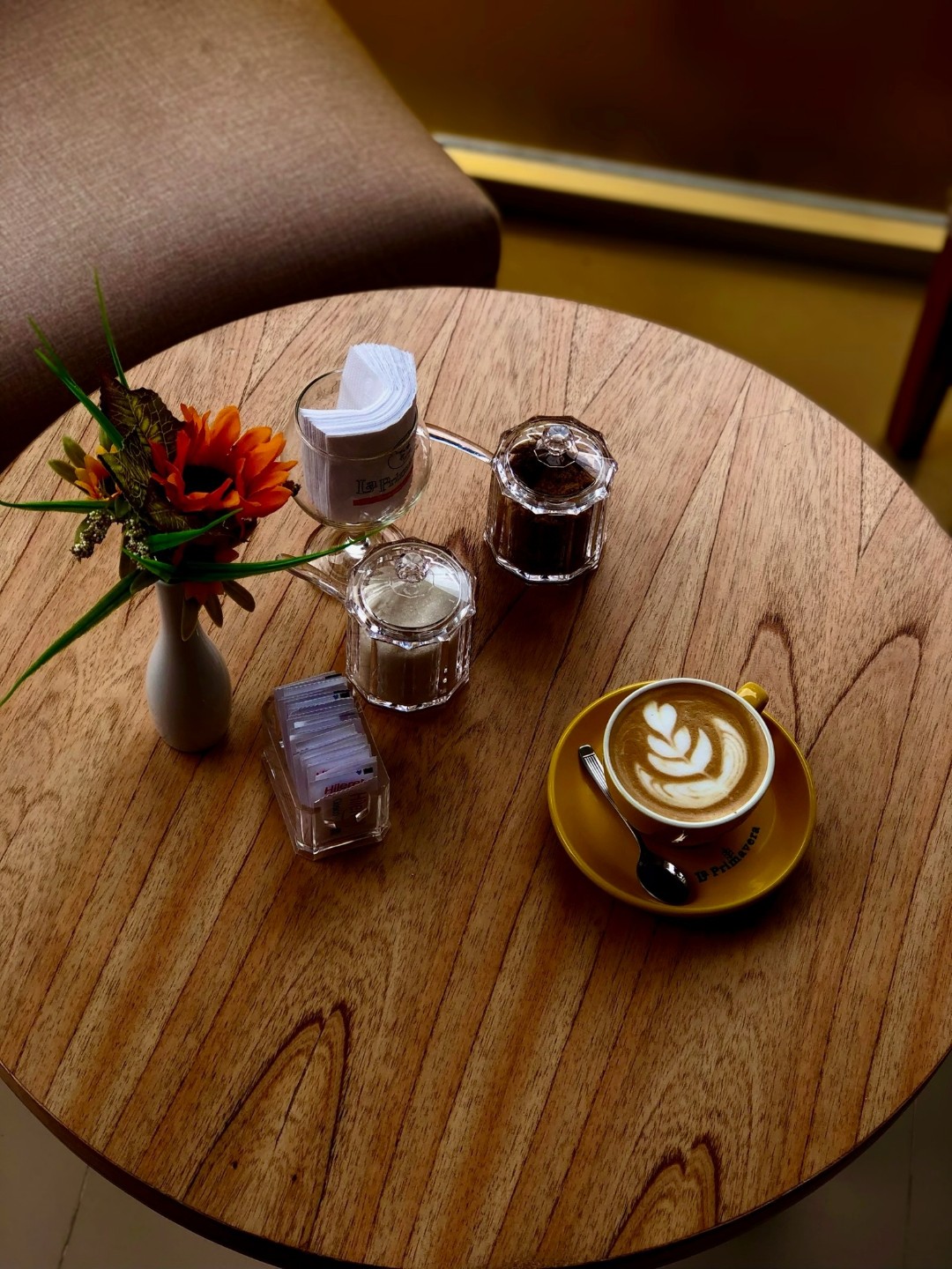 Vista desde arriba de una mesa redonda de madera con un latte con latte art, azucareras de vidrio y un florero con flores naranjas